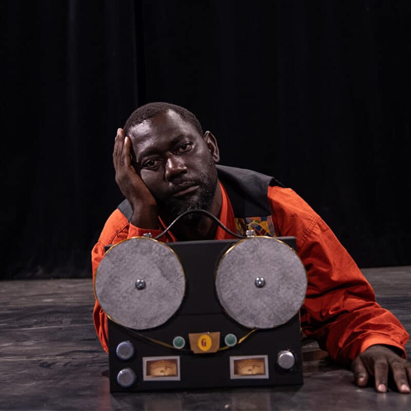 Un homme est allongé sur le ventre. Il tien sa tête avec la main droite, pensif. Devant lui, une sorte de poste radio.