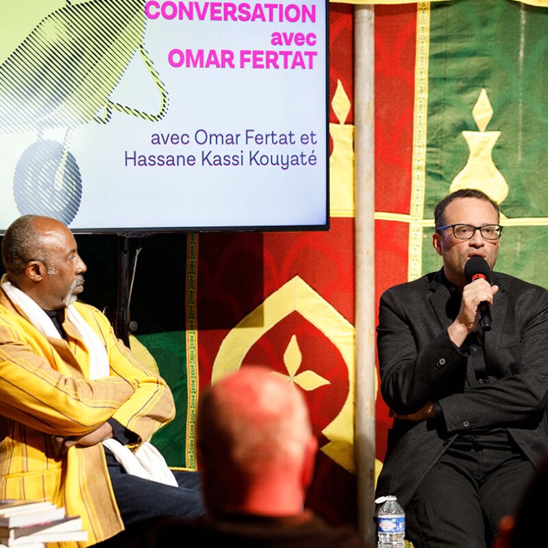 Photo de la rencontre Conversation avec Omar Fertat. À droite on voit Omar Fertat en train de parler dans un micro. À gauche Hassane Kassi Kouyaté, directeur des Francophonies - Des écritures à la scène le regarde. Sur une table à sa droite, une pile de livre est présente. Une télévision est suspendu derrière eux. Il est inscrit 