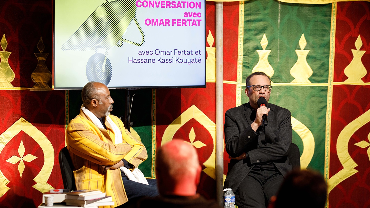Photo de la rencontre Conversation avec Omar Fertat. À droite on voit Omar Fertat en train de parler dans un micro. À gauche Hassane Kassi Kouyaté, directeur des Francophonies - Des écritures à la scène le regarde. Sur une table à sa droite, une pile de livre est présente. Une télévision est suspendu derrière eux. Il est inscrit 