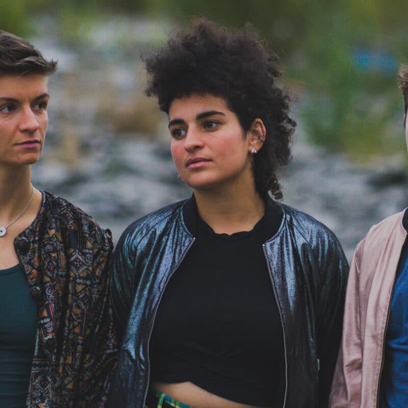 Photo des trois chanteuses du groupe Diluvienne, haut du corps. Paysage de nature en fond. De gauche à droite, Orion Socquet, Nourelaïn Khaoua et Marine Louarn