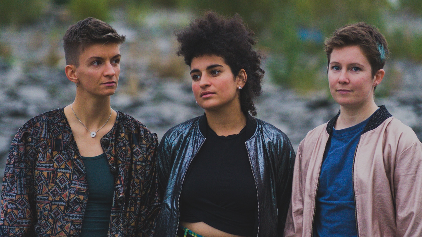 Photo des trois chanteuses du groupe Diluvienne, haut du corps. Paysage de nature en fond. De gauche à droite, Orion Socquet, Nourelaïn Khaoua et Marine Louarn