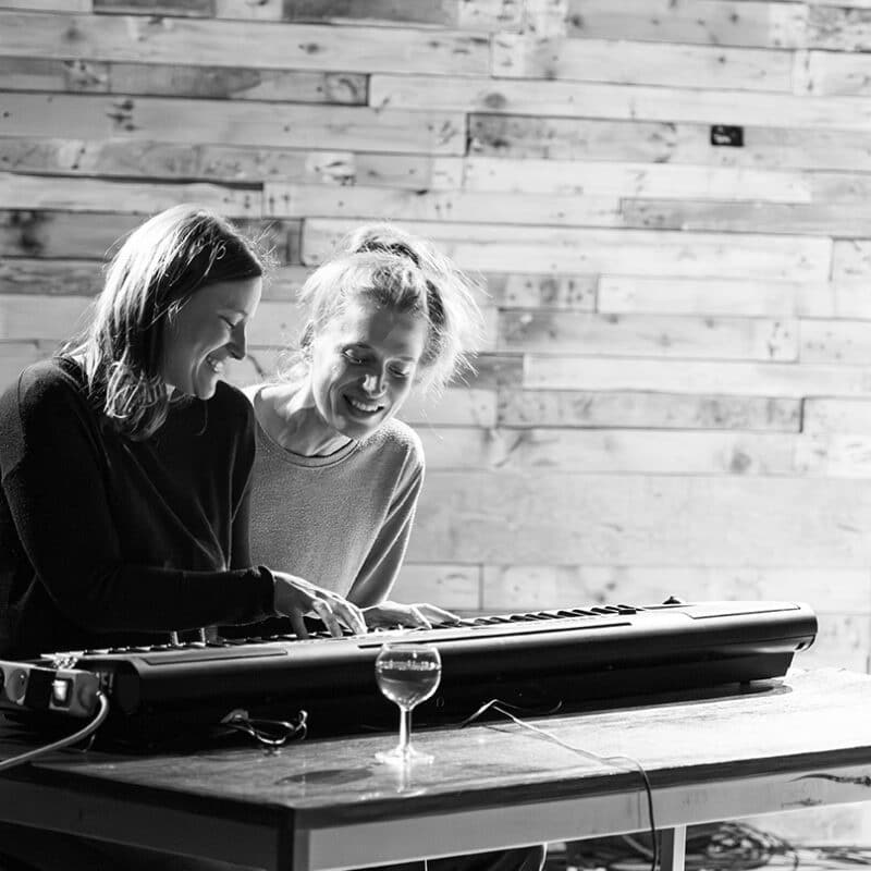 2 femmes rigole derrière un piano. L'une est en train de jouer dessus. Devant le clavier se trouve 2 verres de vins pleins.