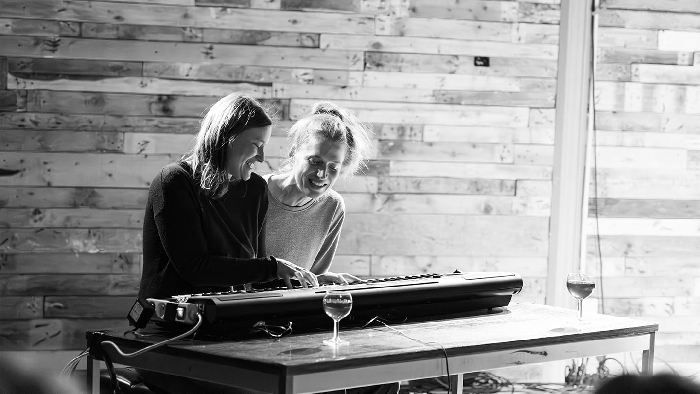 2 femmes rigole derrière un piano. L'une est en train de jouer dessus. Devant le clavier se trouve 2 verres de vins pleins.