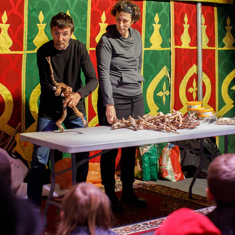 1 personne manipule des bouts de bois pour en faire une marionnette. Un femme à côté le regarde. On distingue des enfants de dos qui le regarde.