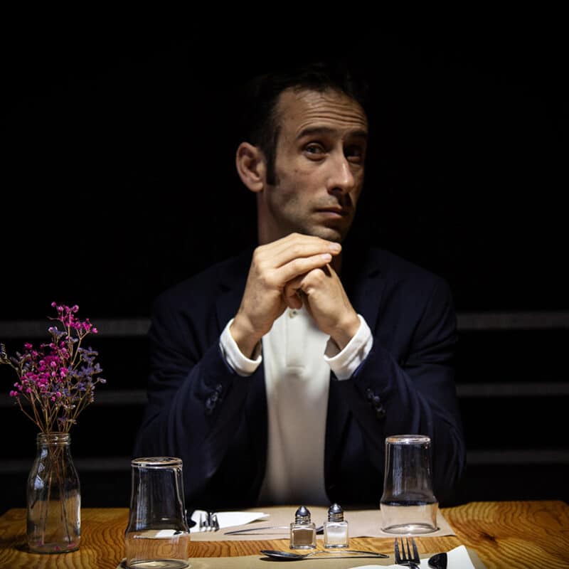 La table est mise pour 2 personnes, comme au restaurant. Un bouquet de fleurs séchées est gauche. Jérôme Pinel est assis à table et regarde en face de lui.