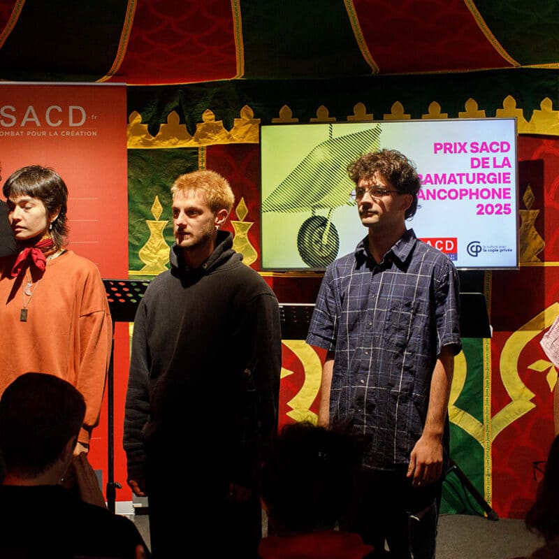 4 comédiens et comédiennes sont face aux publics. Un autre comédien, derrière un pupitre est en train de lire. En arrière plan, on lit Prix SACD de la dramaturgie francophone 2025.