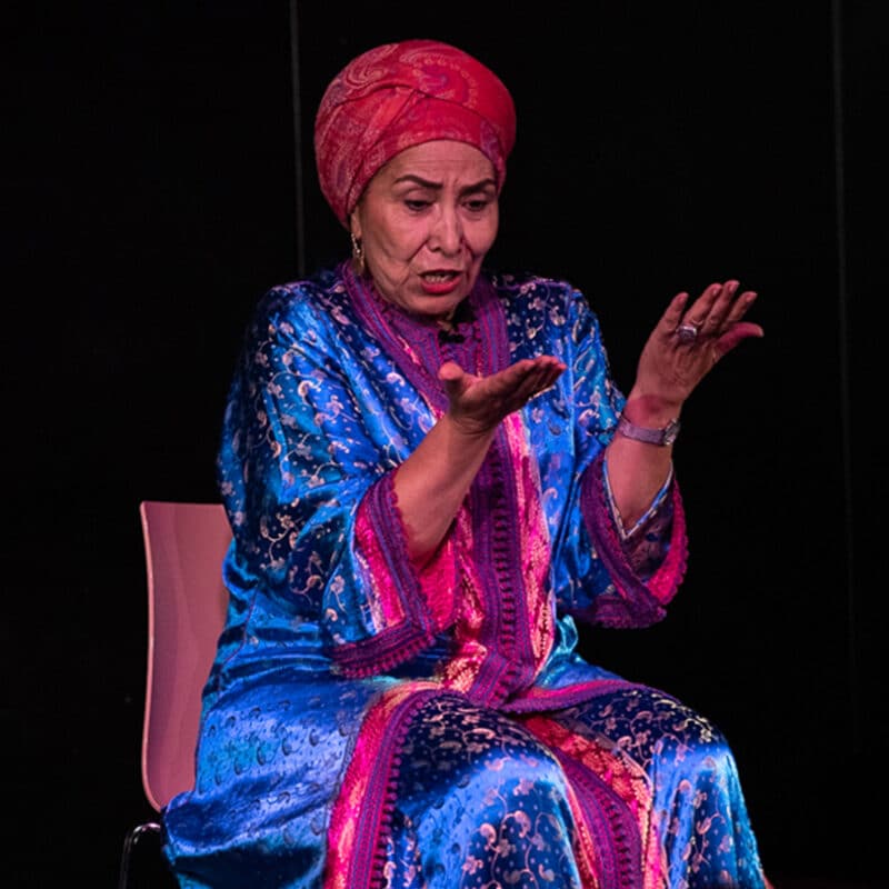 Halima Handame est au centre d'une scène en train de conter. Elle regarde ses mains.
