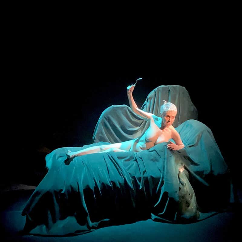 Sur un divan recouvert par des draps bleus, une femme âgée est alongée. elle est nue, une fourchette dans la main gauche qu'elle lève en regardant devant elle.
