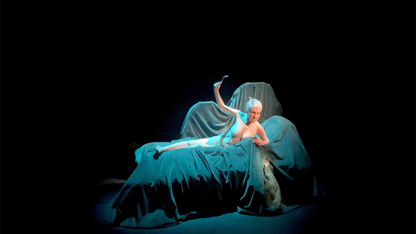 Sur un divan recouvert par des draps bleus, une femme âgée est alongée. elle est nue, une fourchette dans la main gauche qu'elle lève en regardant devant elle.