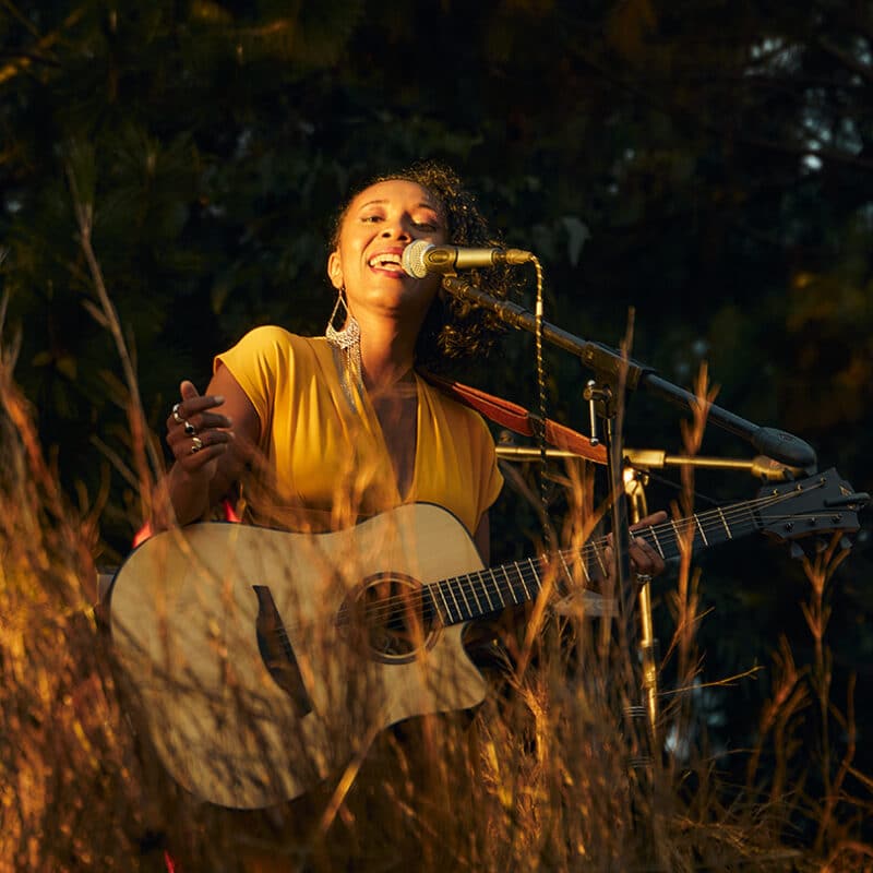 Dina Mialinelina chante, une guitare acoustique à la main. Elle est dans la nature : on voit des arbres en arrière plan et des herbes devant elle.