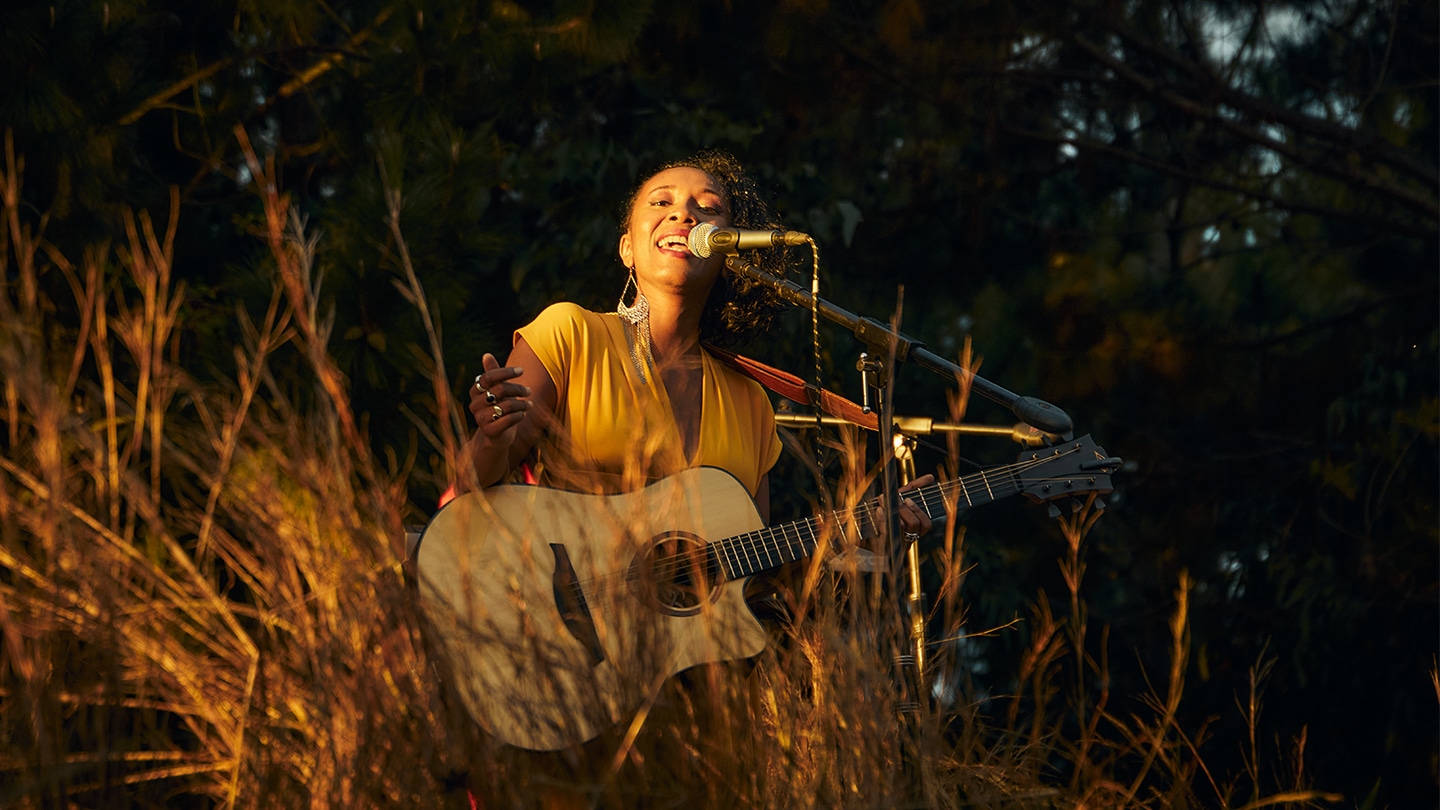 Dina Mialinelina chante, une guitare acoustique à la main. Elle est dans la nature : on voit des arbres en arrière plan et des herbes devant elle.
