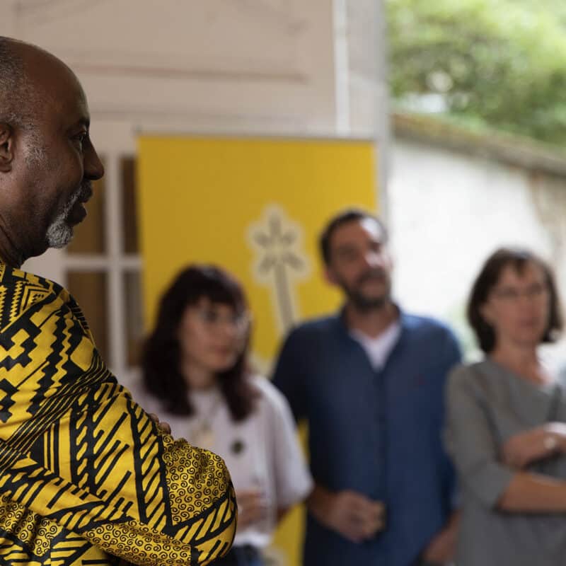 Photo du vernissage de l'exposition Rouge Tapis. Hassane Kassi Kouyaté, directeur des Francophonies - Des écritures à la scène écoute en regardant quelque chose hors champs. En fond, on retrouve 3 personnes de l'équipe des Francophonies.
