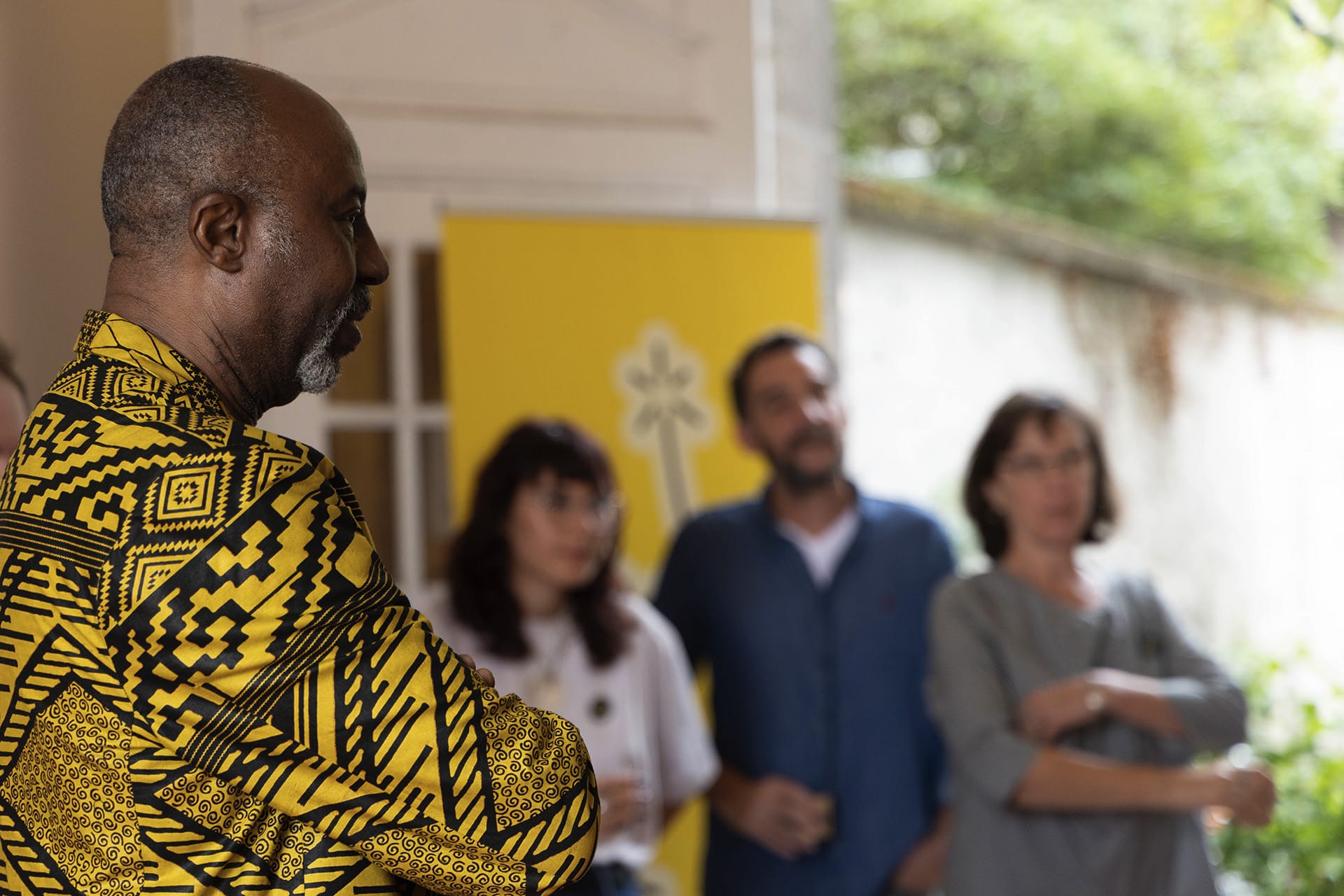 Photo du vernissage de l'exposition Rouge Tapis. Hassane Kassi Kouyaté, directeur des Francophonies - Des écritures à la scène écoute en regardant quelque chose hors champs. En fond, on retrouve 3 personnes de l'équipe des Francophonies.