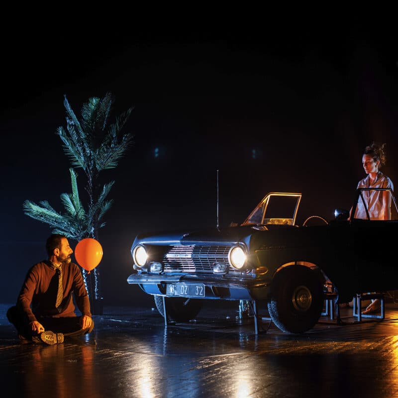 Photo du spectacle .REKORD. Sur scène, un homme est assis en tailleur. Un ballon de baudruche rouche est à côté de sa tête. L'Homme regarde une femme dans une veille Opel Rekord dans la pénombre L'homme est éclairé par les phares de la voiture.