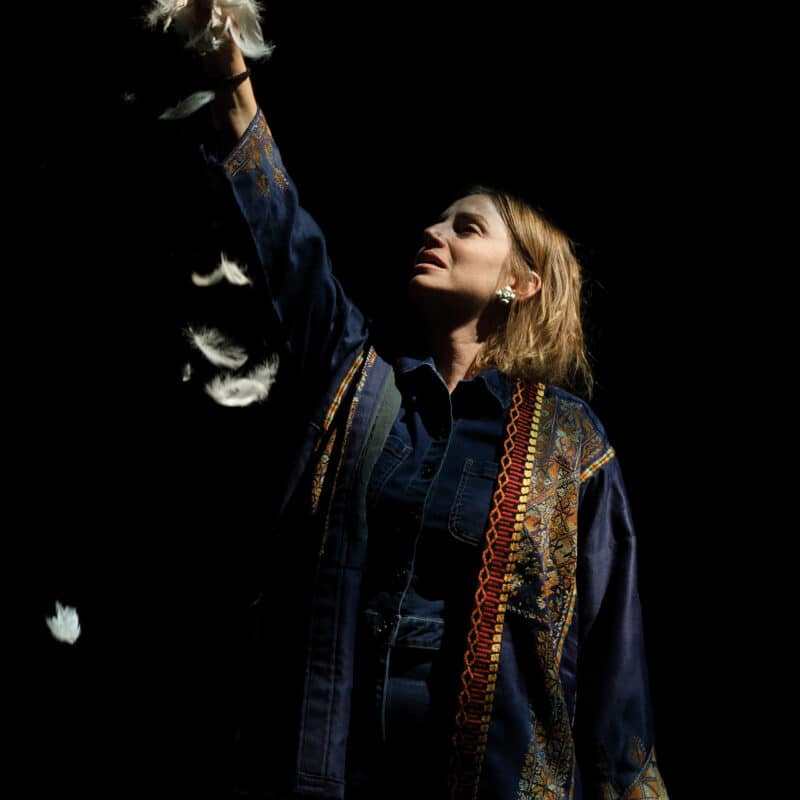 Photo du spectacle .REKORD. Sumaya, une jeune femme tient une poignée de plumes blanches dans sa main droite levée vers le ciel. Elle regarde sa main d'un air triste.