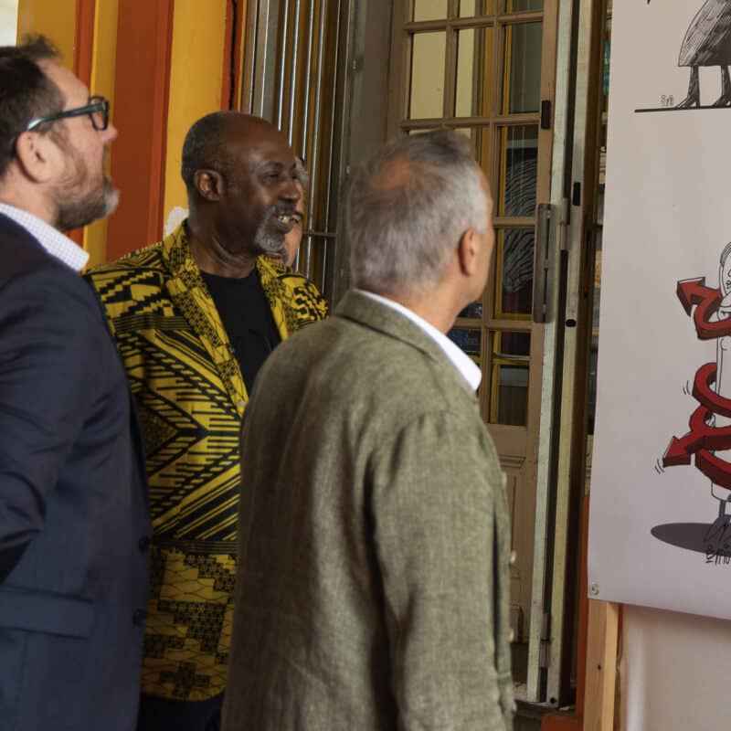 Photo du vernissage de l'exposition Rouge Tapis. 3 hommes regardent un dessin de l'artiste Firoozeh Mozaffari. On y voit une femme voilée dessinée prie au piège par 4 symboles 