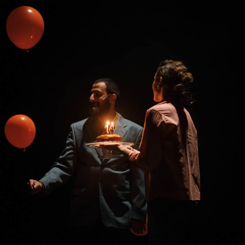Photo du spectacle .REKORD. Un homme face à nous tient dans sa main droite 2 ballons de baudruche rouge : un petit, proche de sa main et un, un peu plus grand, plus en hauteur. Une femme le regarde avec un gâteau d'anniversaire dans les mains avec des bougies 