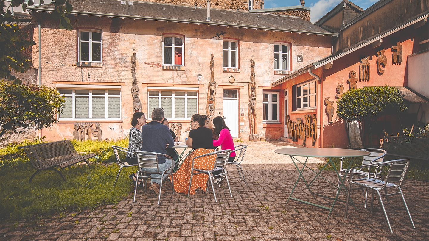 En arrière plan, la Maison des auteurice. Devant, autour d'une table un groupe de personnes discutent.