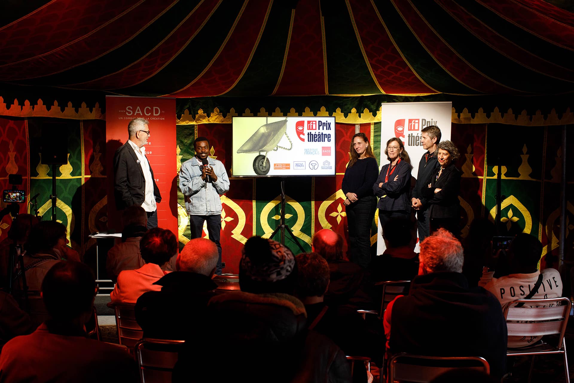 Photo de la remise du Prix RFI Théâtre 2025. À gauche de l'image, Pascal Paradou (journaliste de RFI) regare Israël Nzila (lauréat du Prix) en train de parler. Un groupe de personne le regarde à droite. On voit le public de dos au premier plan.
