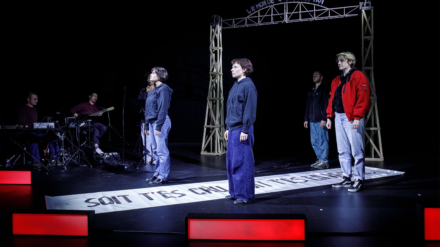 4 jeunes sont debout, regardant au loin, de par et d'autre d'une banderole où il est inscrit 