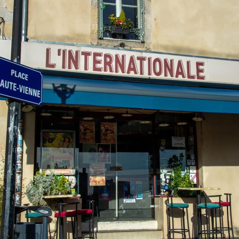 Vue de l'entrée et de la terrasse du bar restaurant L'Internationale avec quelques tabourets et tables hautes et le panneau du nom de la place : Haute-Vienne