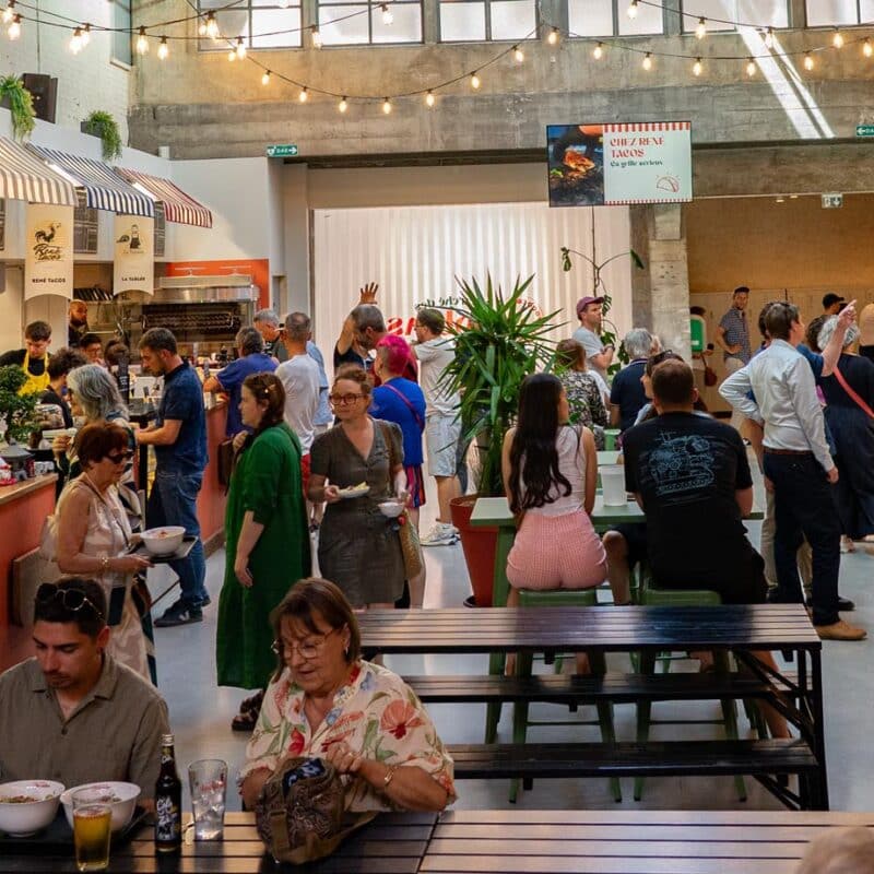 Photo de l'intérieur du marché des Jacobins avec des publics assis à des tables hautes et des tables avec des bancs au milieu et de chaque côté des stands des différents commerces de nourriture
