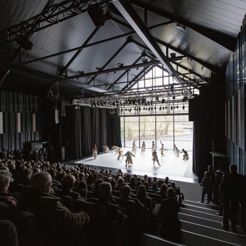 Photo de la scène de l'auditorium depuis la haut du gradin. Il y a du public dans le gradin et sur scène, il y a plusieurs danseurs et danseuses. Les rideaux du fond de scène sont ouverts et permettent de voir à l'extérieur du théâtre