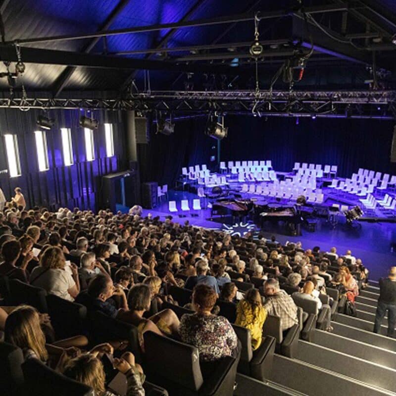 Photo de l'auditorium Sophie Dessus depuis le haut du gradin. Il y a du public et sur scène sont installés des chaises disposées pour un orchestre et deux pianos à queue face à face en front de scène