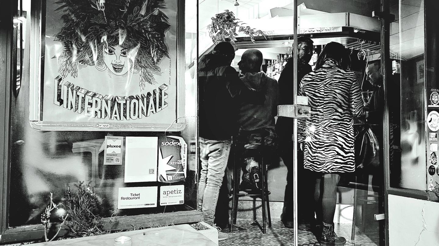 Photo représentant la devanture du bar-restaurant L'Internationale. On peut voir à travers la vitrine des personnes accoudées au bar dont une personne avec un manteau au motif de zèbre.