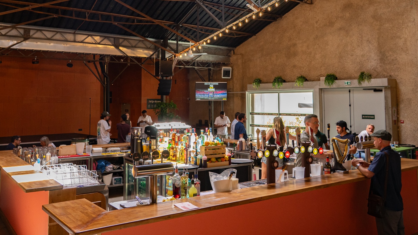 Photo de l'intérieur du marché des Jacobins où on peut voir le bar central carré, des personnes qui servent et des personnes accoudées au bar qui commandent. Dans le fond, on voit un écran et des personnes qui discutent debout.