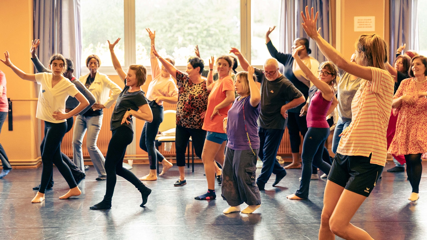 Un groupe d'amateurs et amatrices danse. Ils lèvent la main guidés par une jeune femme de dos.