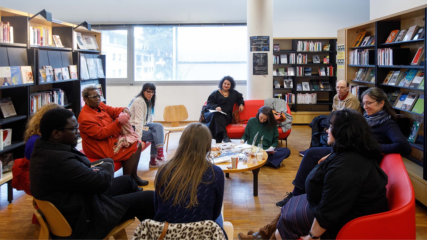 Dans une bibliothèque, un groupe de personnes est en cercle et discute. Au milieu, une table basse avec des livres.