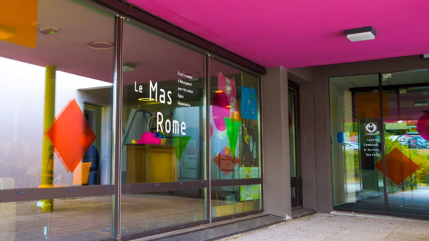 Photo de l'entrée de l'EHPAD du Mas Rome, avec des portes vitrées sur lesquelles est écrit le nom de l'établissement, et des couleurs flachies